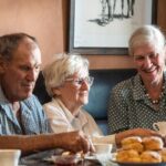 Three older adults sit at a table with tea and scones, smiling and enjoying each other’s company in a warmly lit room.