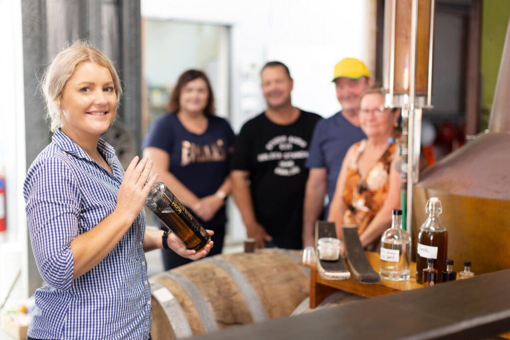 A woman holds a bottle and smiles at the camera in a distillery, while four adults stand blurred in the background near barrels and distilling equipment.