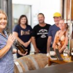 A woman holds a bottle and smiles at the camera in a distillery, while four adults stand blurred in the background near barrels and distilling equipment.