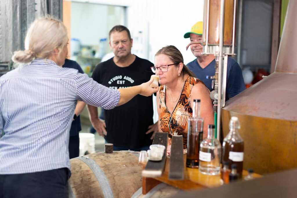 A group of adults observe as a woman smells the contents of a small jar in a distillery, surrounded by bottles and equipment.