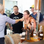 A group of adults observe as a woman smells the contents of a small jar in a distillery, surrounded by bottles and equipment.