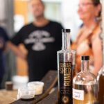 A bottle of Sugar Shed Rum and a clear labeled bottle sit on a counter with tasting samples; two people are out of focus in the background.