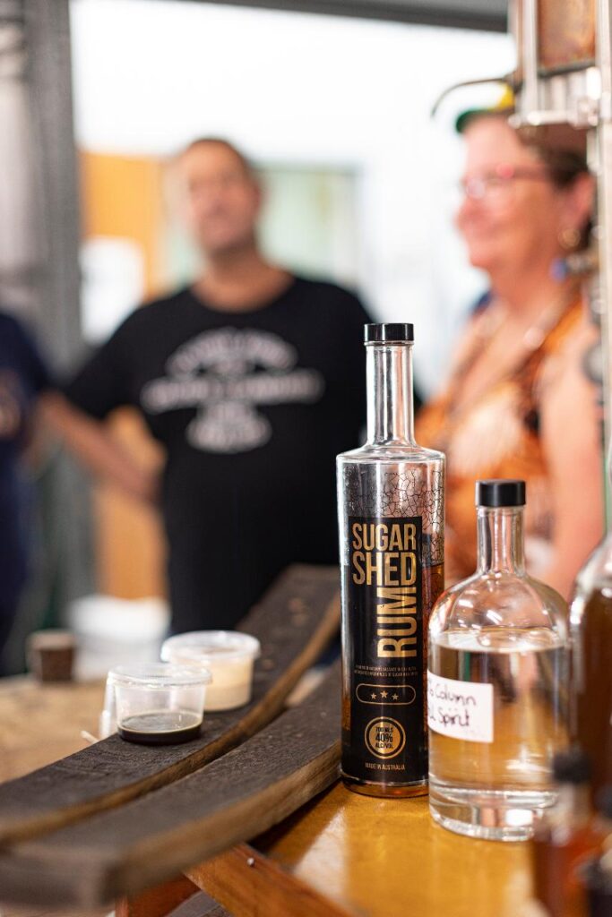 A bottle of Sugar Shed Rum and a clear labeled bottle sit on a counter with tasting samples; two people are out of focus in the background.