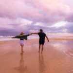 A couple walks hand-in-hand on a wet sandy beach under a cloudy, pinkish sky.