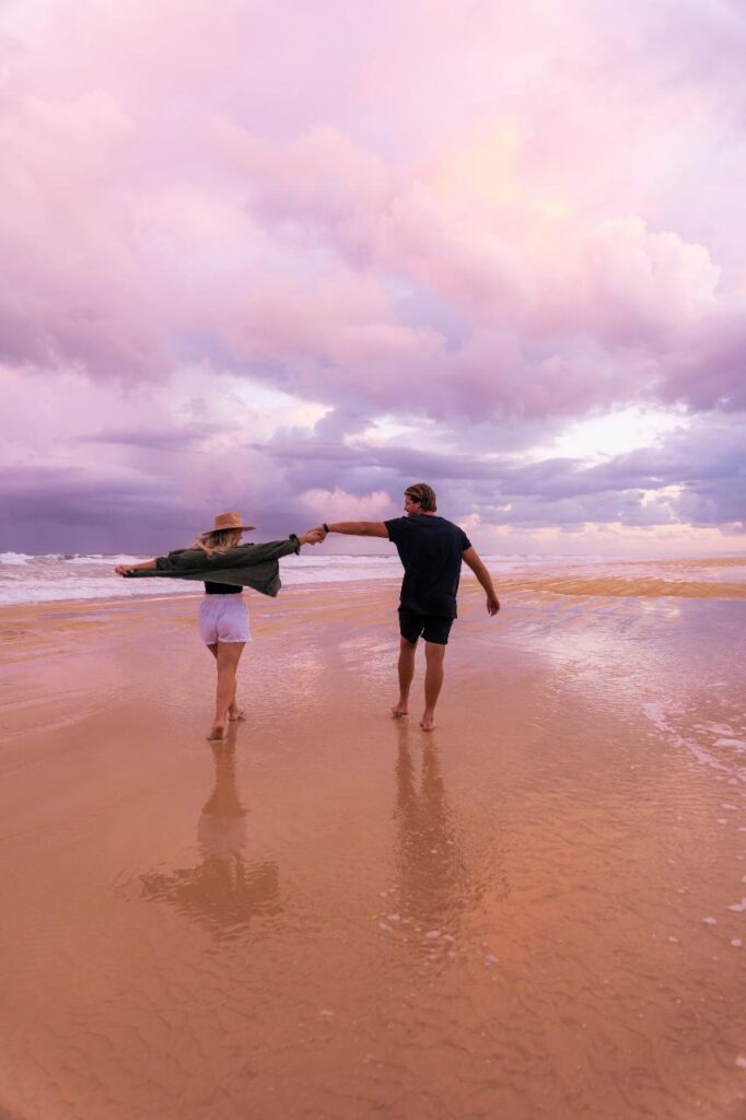 A couple walks hand-in-hand on a wet sandy beach under a cloudy, pinkish sky.