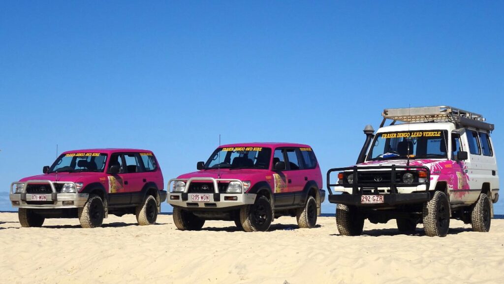 Three off-road vehicles, two pink and one white with pink accents, are parked side by side on sandy terrain under a clear blue sky.