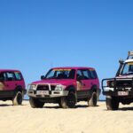 Three off-road vehicles, two pink and one white with pink accents, are parked side by side on sandy terrain under a clear blue sky.