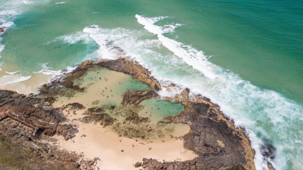 Aerial view of a rocky coastal tide pool with people swimming, surrounded by sandy beach and turquoise ocean waves.