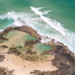 Aerial view of a rocky coastal tide pool with people swimming, surrounded by sandy beach and turquoise ocean waves.