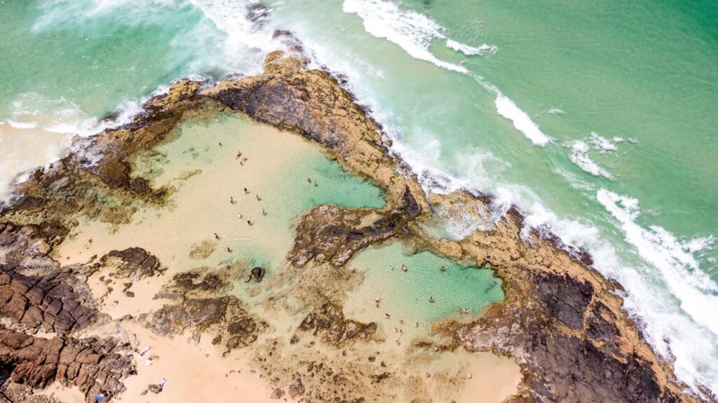 Aerial view of a rocky coastline with natural tidal pools and people swimming; ocean waves approach the shore.