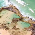 Aerial view of a rocky coastline with natural tidal pools and people swimming; ocean waves approach the shore.