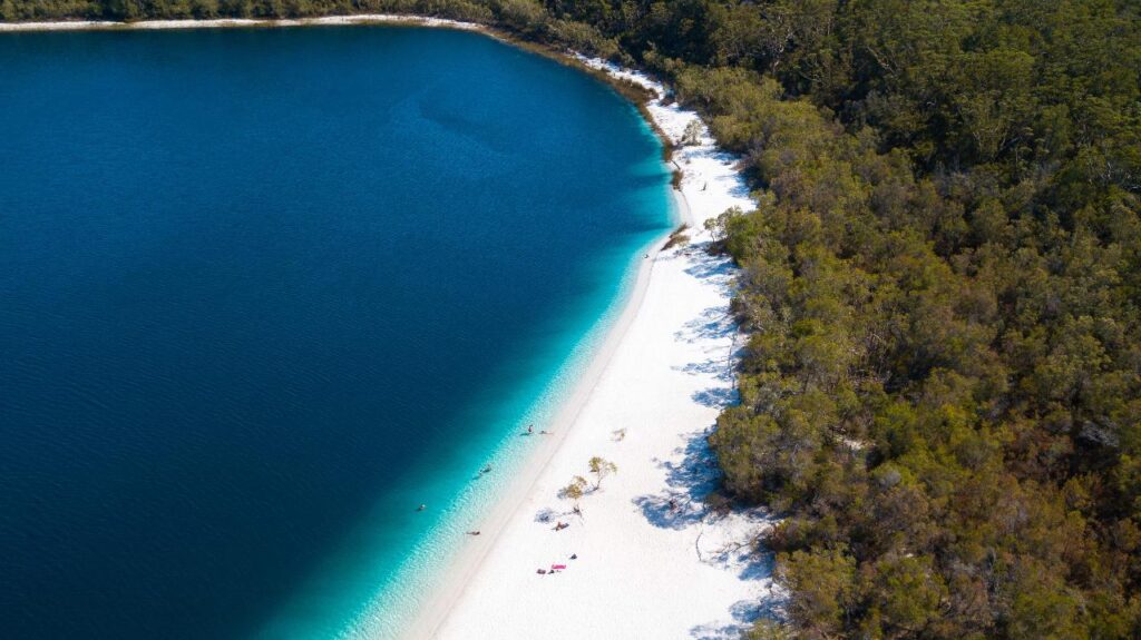 Aerial view of a lake with deep blue water, white sandy beach, and dense green forest along the shoreline. People are visible relaxing on the beach.