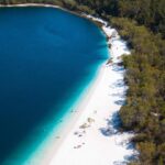 Aerial view of a lake with deep blue water, white sandy beach, and dense green forest along the shoreline. People are visible relaxing on the beach.