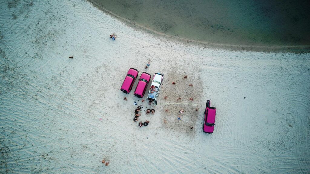 Aerial view of four vehicles parked on a sandy beach with people gathered nearby and along the shoreline.