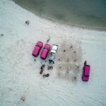 Aerial view of four vehicles parked on a sandy beach with people gathered nearby and along the shoreline.