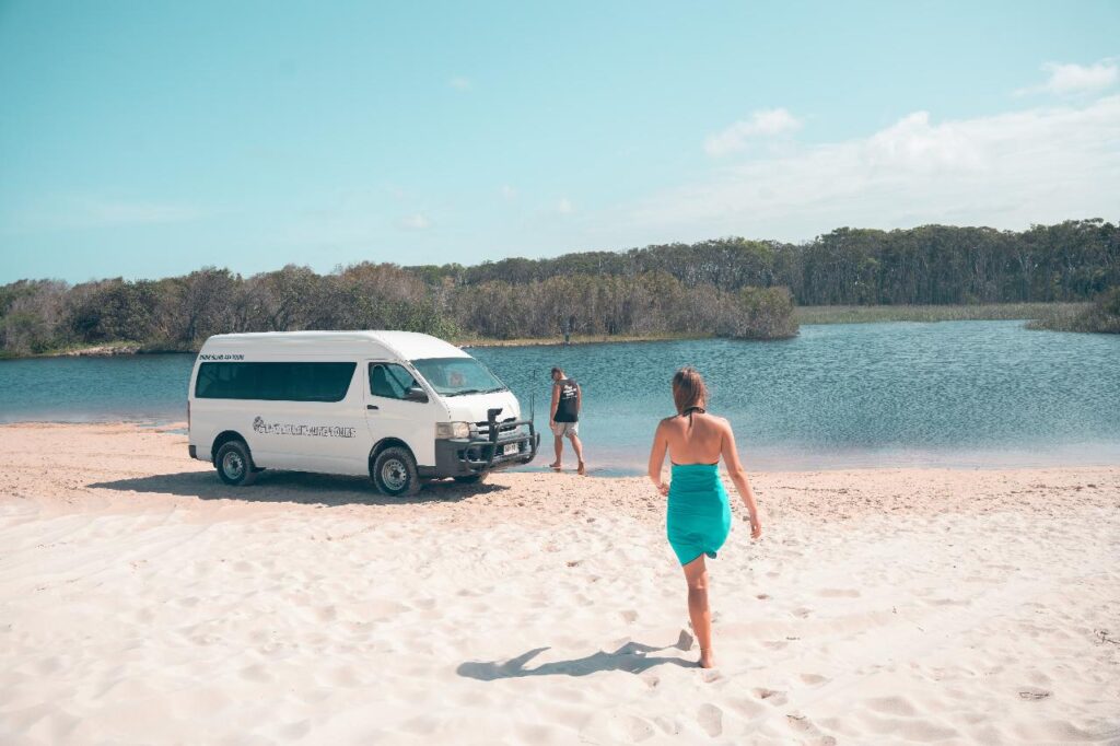 A woman in a turquoise dress walks toward a white van and another person standing by a lakeshore, with sand, water, and trees in the background under a clear sky.