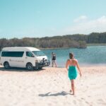 A woman in a turquoise dress walks toward a white van and another person standing by a lakeshore, with sand, water, and trees in the background under a clear sky.