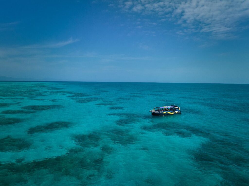 A small boat floats on clear blue ocean water under a partly cloudy sky, with coral reefs visible beneath the surface.