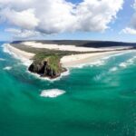 Aerial view of a rocky headland surrounded by turquoise ocean, white sandy beaches, and a stretch of coastal dunes under a partly cloudy sky.