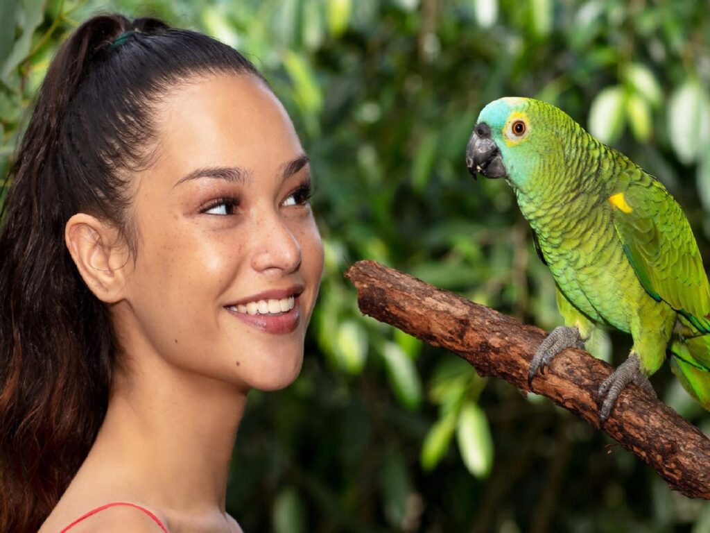 A woman with a ponytail smiles at a green parrot perched on a branch, with green foliage in the background.