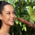 A woman with a ponytail smiles at a green parrot perched on a branch, with green foliage in the background.