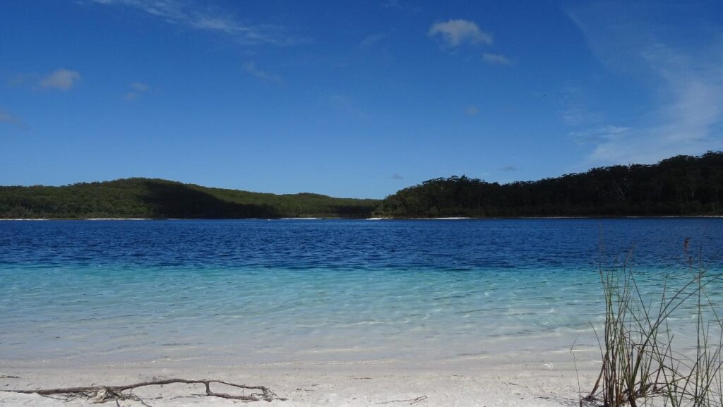 A clear blue lake with calm water, white sandy shore in the foreground, and forested hills in the background under a blue sky.
