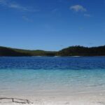 A clear blue lake with calm water, white sandy shore in the foreground, and forested hills in the background under a blue sky.