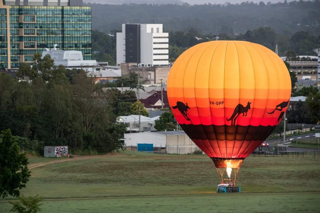 A hot air balloon with kangaroo silhouettes is inflated on a grassy field near city buildings, early in the morning.