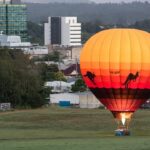 A hot air balloon with kangaroo silhouettes is inflated on a grassy field near city buildings, early in the morning.