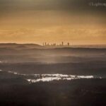 A hazy landscape with layered hills in the foreground and a distant city skyline under a cloudy, golden sky.