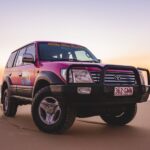 A pink Toyota Land Cruiser with off-road tires is parked on a sandy beach at sunset, with the ocean and colorful sky in the background.