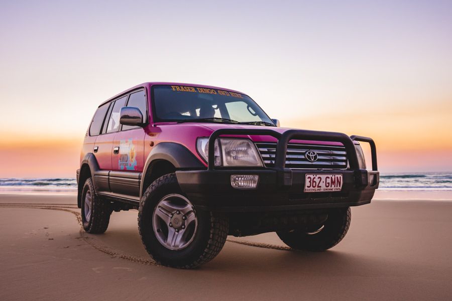 A pink Toyota Land Cruiser with off-road tires is parked on a sandy beach at sunset, with the ocean and colorful sky in the background.