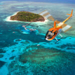A bright orange helicopter flies over clear blue water near a small, lush island with boats anchored nearby.