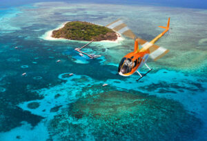 A bright orange helicopter flies over clear blue water near a small, lush island with boats anchored nearby.