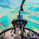View from inside a helicopter cockpit flying over turquoise waters and coral reefs, showing the control panel and pilot’s feet.