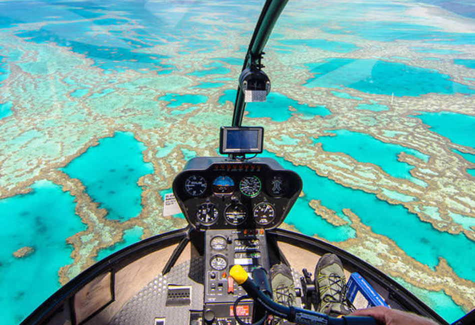 View from inside a helicopter cockpit flying over turquoise waters and coral reefs, showing the control panel and pilot’s feet.