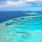 Aerial view of a coral reef in clear turquoise water, with the deep blue ocean and a partly cloudy sky in the background.