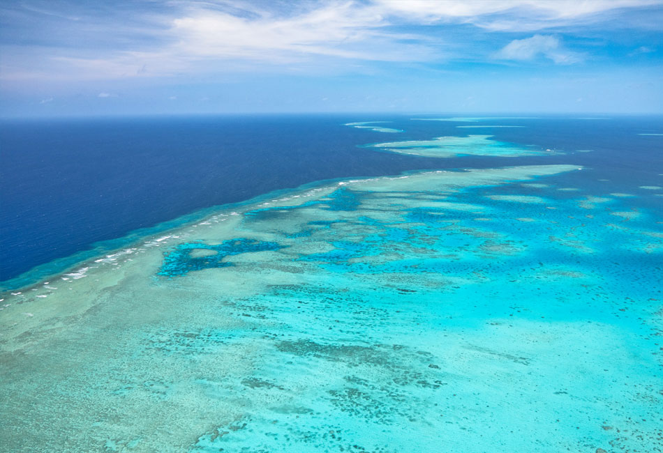 Aerial view of a coral reef in clear turquoise water, with the deep blue ocean and a partly cloudy sky in the background.