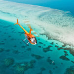An orange helicopter flies over clear blue water and coral reefs near a sandy coastline.