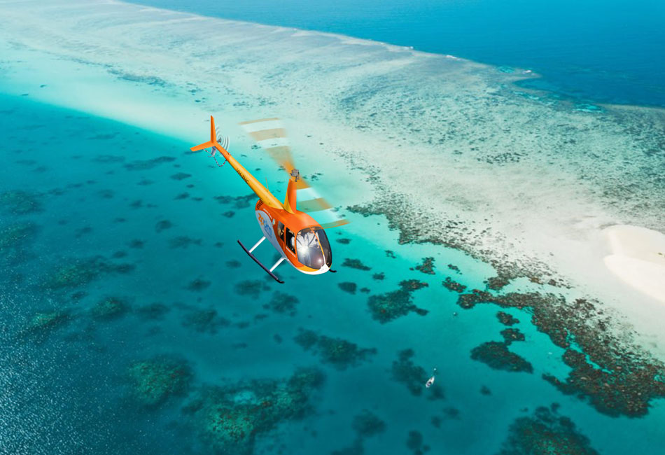 An orange helicopter flies over clear blue water and coral reefs near a sandy coastline.