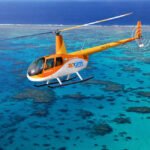 An orange and white helicopter with "Zoom Helicopters" branding flies over clear blue ocean water and coral reefs.