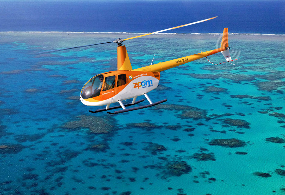 An orange and white helicopter with "Zoom Helicopters" branding flies over clear blue ocean water and coral reefs.