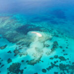 Aerial view of a small sandbar surrounded by turquoise water and coral reefs in a clear blue ocean.