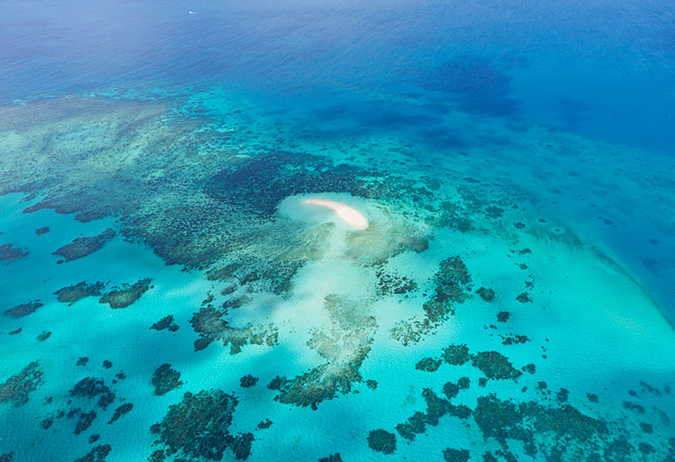 Aerial view of a small sandbar surrounded by turquoise water and coral reefs in a clear blue ocean.