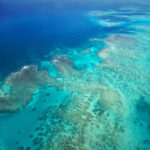 Aerial view of turquoise waters and coral formations in a large, shallow reef area extending into the deep blue ocean.