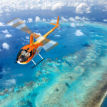 An orange helicopter flies over a vibrant blue ocean with coral reefs and scattered clouds below.
