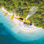 A yellow helicopter flies above a sandy beach with turquoise water and dense green trees, viewed from above.