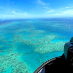 Aerial view from a helicopter cockpit overlooking turquoise waters and coral formations, likely part of a large reef system under a clear blue sky.
