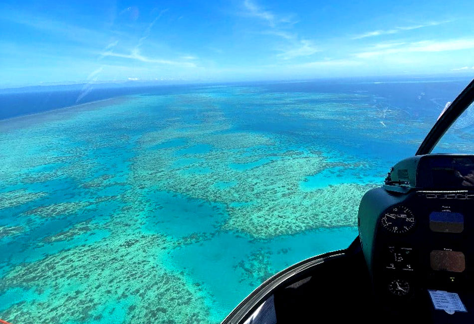 Aerial view from a helicopter cockpit overlooking turquoise waters and coral formations, likely part of a large reef system under a clear blue sky.