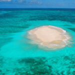 A small sandy island surrounded by clear turquoise ocean water under a blue sky.